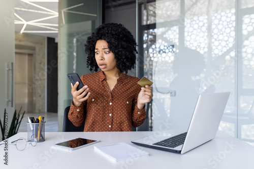 Φωτογραφία A shocked young Black woman at her office desk, looking perplexedly at her smartphone while holding a credit card, possibly dealing with an unexpected transaction