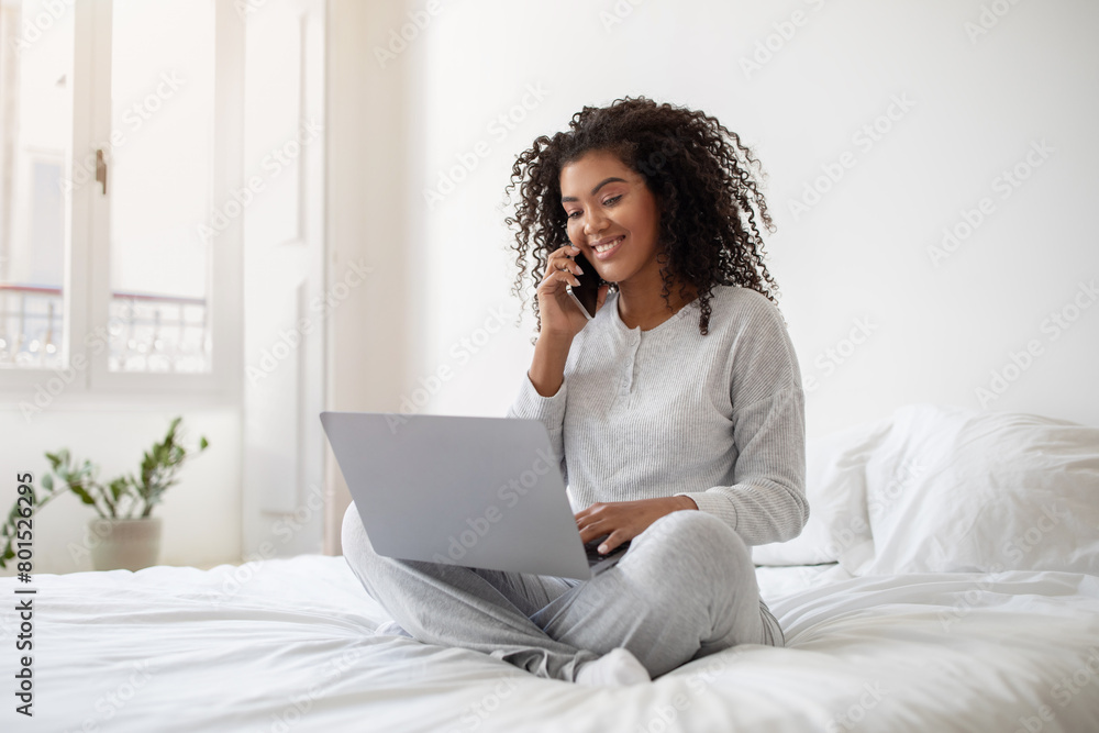 Hispanic woman is sitting on a bed, multitasking by talking on her cell phone while using a laptop. She appears focused and engaged in her activities, balancing both devices comfortably. Hispanic woman is sitting on a bed, multitasking by talking on her cell phone while using a laptop. She appears focused and engaged in her activities, balancing both devices comfortably.