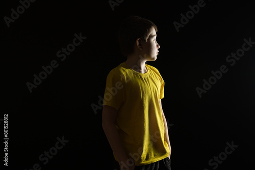 A young boy in a yellow shirt stands profile against a black background, with half of his face illuminated. © Sergey