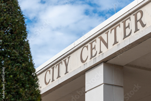 A city center sign on the exterior of a cream colored building with an overhang and square column. The recreation facility has a large green hedge in front with a blue sky in the background.