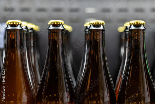 Multiple rows of long sleek neck brown beer bottles. The craft beer bottles are filled with a cold ale and capped with gold crowned caps. The liquor is stored in a metal cooler in rows with no labels