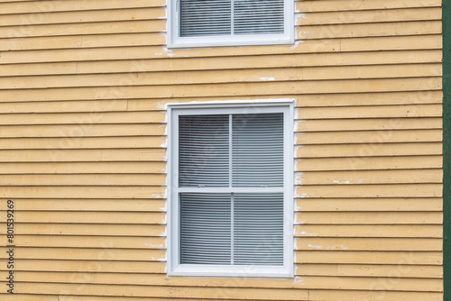 The exterior wall has a vibrant yellow wooden clapboard siding wall with a double-hung window. The wide colonial trim around the window is white. There are small white blinds in the window. 