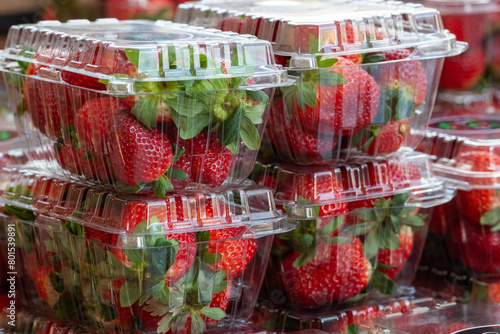 Clear plastic containers of fresh red, ripe strawberries with the stems still on them. The packages are stacked on top of each other on a grocery store shelf.  The berries are firm, rich, and vibrant.
