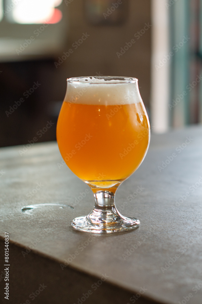 A small chalice sample glass of pale ale craft beer. The liquid has a yellow tint. A clear beer glass sits on the edge of a wooden table at a microbrewery. The background has green garden shrubs. A small chalice sample glass of pale ale craft beer. The liquid has a yellow tint. A clear beer glass sits on the edge of a wooden table at a microbrewery. The background has green garden shrubs.