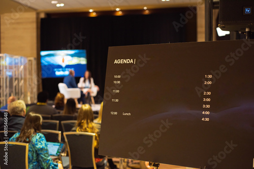 Blank meeting agenda template on telescoping display easel with blurred speakers interview table at business conference convention center in Dallas, Texas, news reporters with laptop memo notes