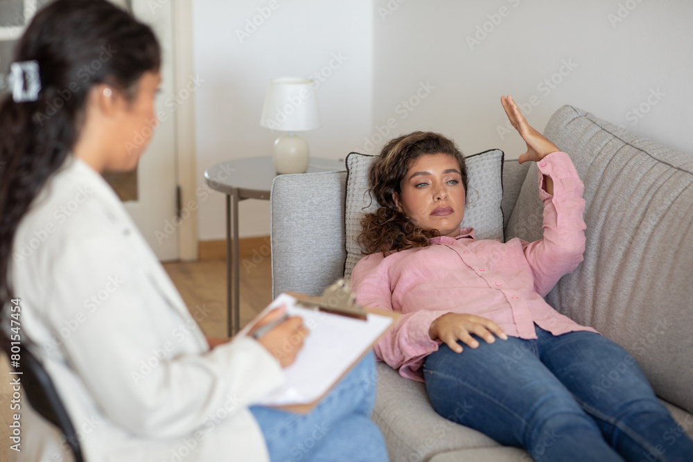 Emotional young woman suffering from anxiety, laying on couch at clinic, sharing her thoughts, have therapeutic session with psychologist Emotional young woman suffering from anxiety, laying on couch at clinic, sharing her thoughts, have therapeutic session with psychologist