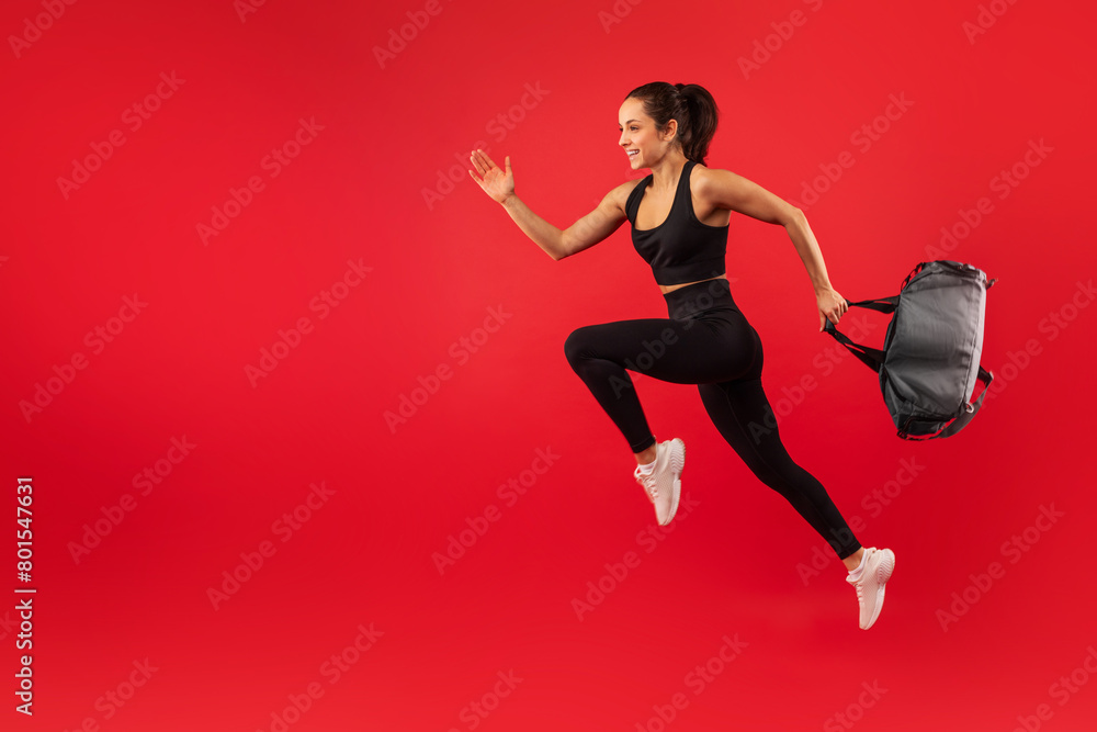 A young woman is captured mid-jump against a vibrant red backdrop, exuding energy and happiness. She is dressed in sporty black attire and white sneakers, with a backpack in one hand