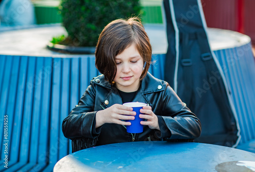 A girl with a square hairstyle, sitting in a summer cafe and drinking tea from a disposable cup, with a guitar in the background