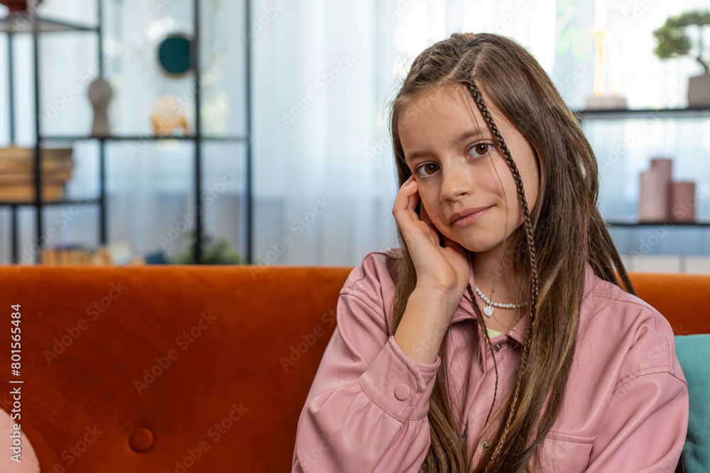 Portrait of happy smiling young girl child sitting on comfy sofa couch looking at camera in living room at home apartment. Cute teenage female school kid with brown long hair. Lifestyles.