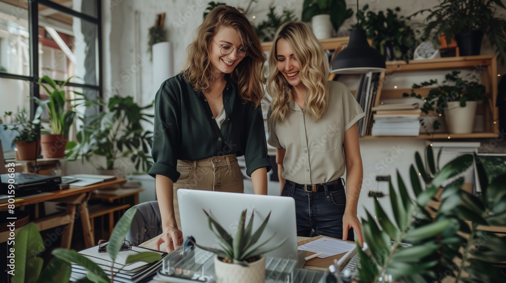 Two smiling professional female partners or coworkers, happy business women entrepreneurs working together in office looking at laptop using computer writing notes standing at work desk. Two smiling professional female partners or coworkers, happy business women entrepreneurs working together in office looking at laptop using computer writing notes standing at work desk.