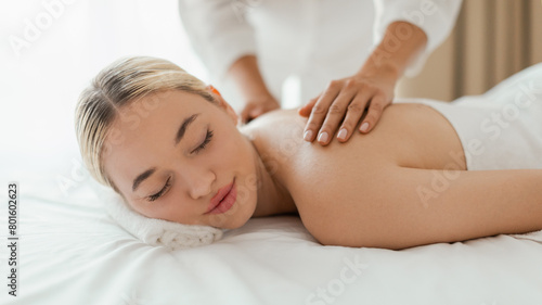 A woman lying face down on a massage table at a spa, receiving a back massage from a massage therapist, panorama