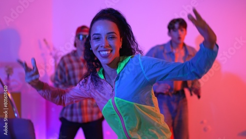 Attractive happy dancer looking at camera while smart woman moving at rhyme with neon light. Professional hispanic performer break dancing while wearing colorful cloth with diverse friend. Regalement. photo