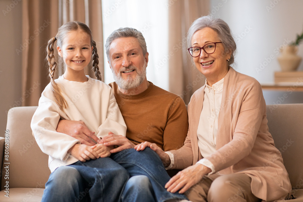 A heartwarming scene captures a young girl with long, braided hair sitting between her grandparents on a beige couch. The elderly couple and the girl are all smiling warmly