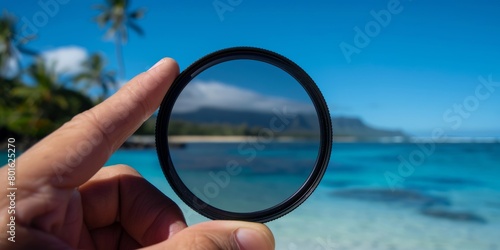 A hand holding a circular polarizing filter in front of the camera, with clear blue sky and island beach background. photo