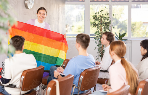 Young teacher showing big transgender flag to group of grown-up student in light classroom