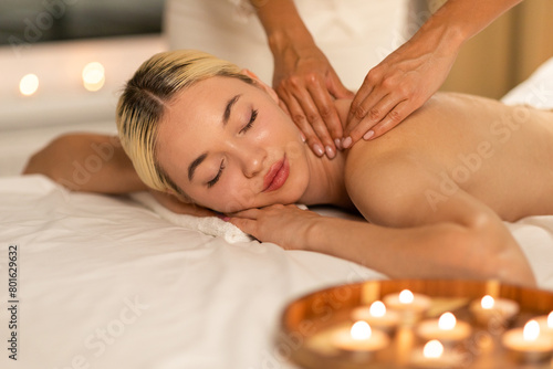 Beautiful woman lies face down on a massage table, receiving a back massage from a professional masseuse in a spa setting. The masseuses hands apply pressure on the woman back