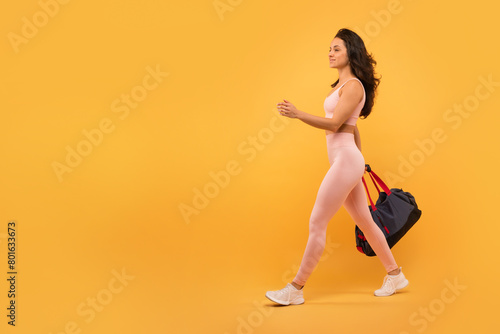A young woman with long hair, dressed in a form-fitting activewear outfit, walks with purpose while carrying a gym bag, isolated on yellow background