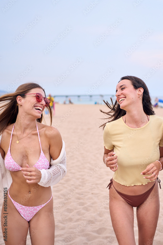 Vertical. Two cheerful young Caucasian women enjoying running having fun on the beach in swimming costume. Female friends having fun in summer vacation together outdoor. Generation z in holidays