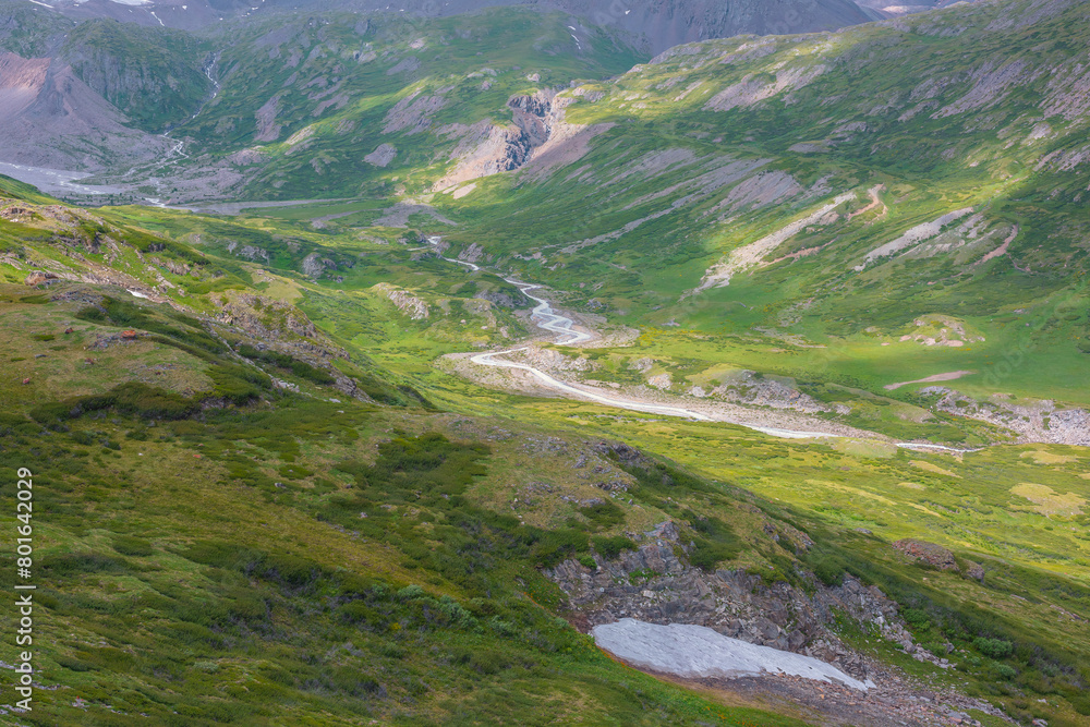 Fototapeta premium Colorful scenery of green alpine valley with snake river among hills and rocks with lush flora in sunlight. Scenic view to serpentine river in mountain valley and shadows of clouds on rich vegetation.