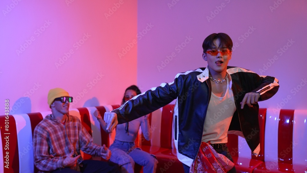 Asian man standing and dancing with pop music while multicultural friend sitting behind at home with led light. Attractive street dancer moving to hip hop music while looking at camera. Regalement.
