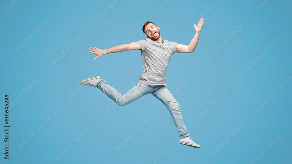 Funny millennial guy jumping in air and fooling on blue background, full length, empty space