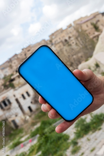 Man hand holds black smartphone with blue screen against the backdrop of a blurred city. (ID: 801667665)