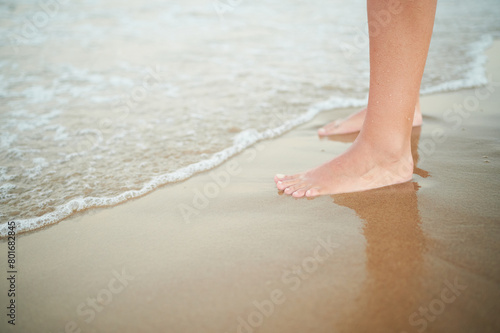 Feet of an unrecognizable woman are bathed by waves of sea water while standing on the shore of the beach.