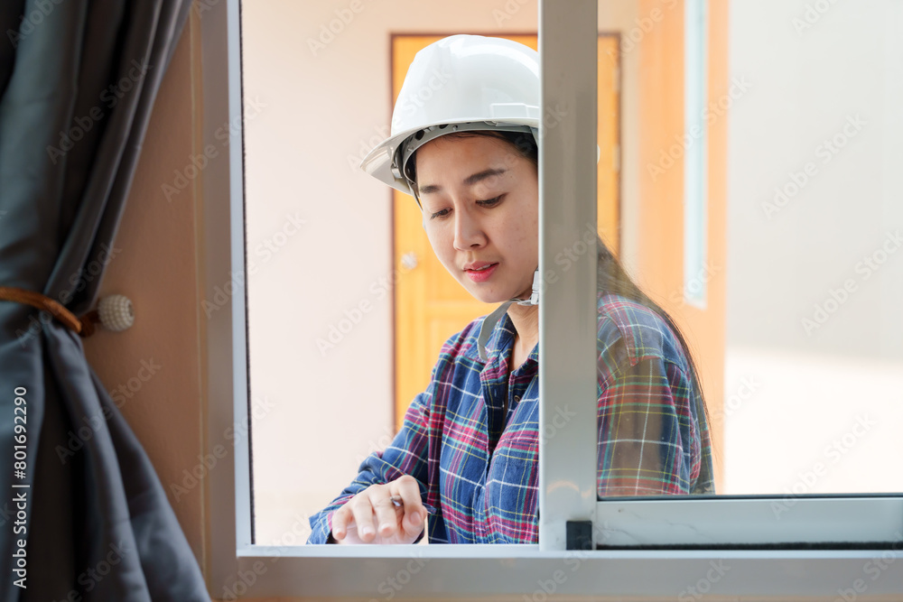 Young Asian female inspector with hard hat writing on clipboard, focusing on window inspection, plaid shirt, safety gear. Inspectors inspect the completed house. Young Asian female inspector with hard hat writing on clipboard, focusing on window inspection, plaid shirt, safety gear. Inspectors inspect the completed house.