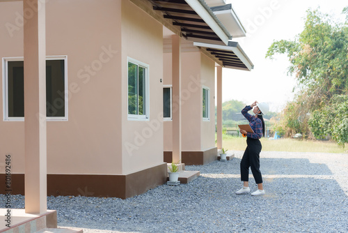 Asian female in safety helmet, gazing upwards, standing outside, clipboard in hand, inspecting newly constructed house. Inspectors inspect the completed house. © Ekkasit A Siam