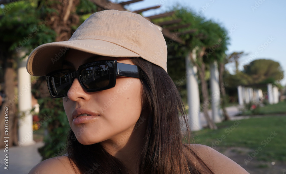Portrait of a young confident woman wearing sunglasses and a cap, in the park at sunset