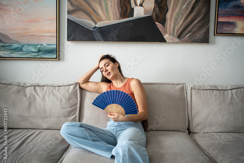 Overheated woman sits on couch in living room at hot summer weather day feel discomfort suffers from heat waving blue fan to cool. Exhausted girl artist sweating cannot working without air conditioner