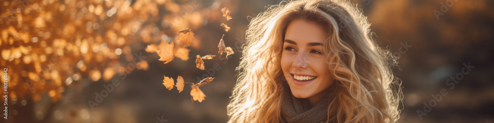 Fototapeta premium A woman with long blonde hair is smiling in front of a tree with leaves falling