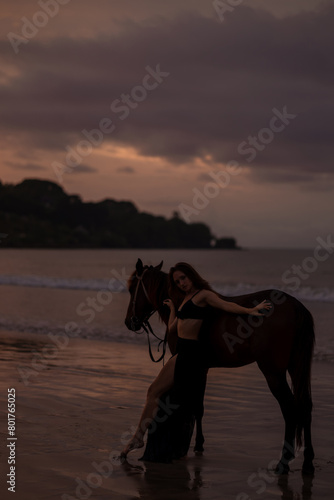 Beautiful woman in black dressnstand near  horse on the beach at sunset, opcean on background photo