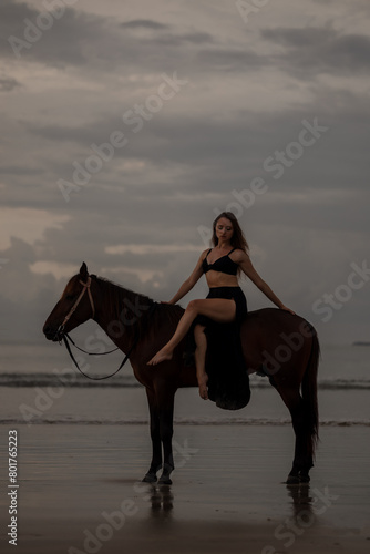 Young long hair woman in black drss is riding horse on seascape background. Rider female drives her horse in nature on evening sunset light background