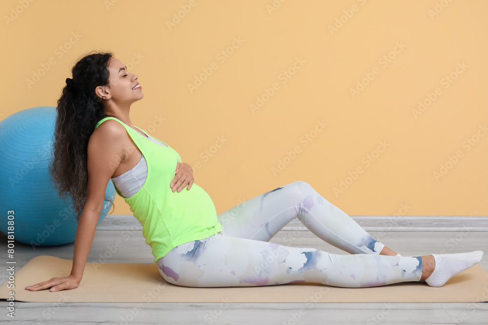 Beautiful young pregnant African-American woman training on yoga mat near yellow wall