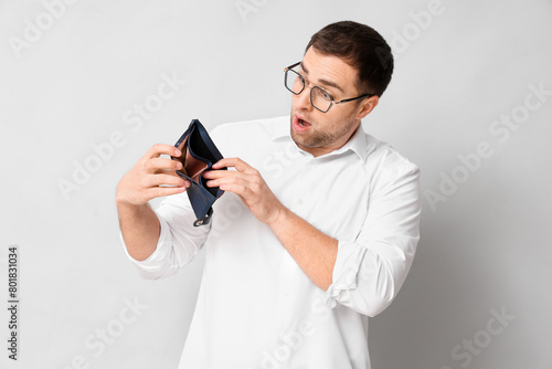 Shocked young man in eyeglasses with empty wallet on white background