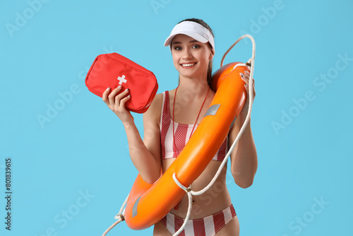 Young female lifeguard in cap with rescue ring and first aid kit on blue background