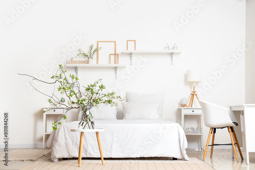 Vase with blooming branches on coffee table near cozy bed in white bedroom