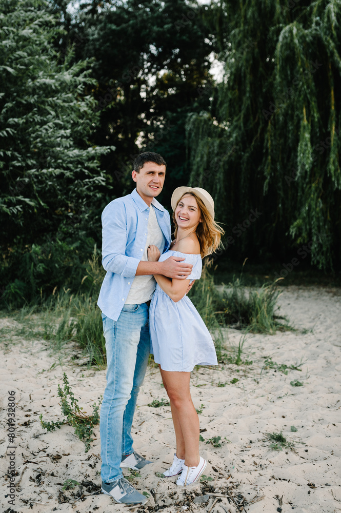 Fototapeta premium Man and woman embrace on sand sea. Happy couple in love hugging and looking at camera on seashore. Female and male standing on beach ocean and enjoying sunny summer day on vacation.