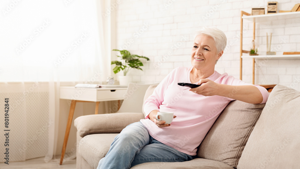 Elderly woman sitting comfortably on a couch, holding a remote control in her hand. She appears to be focused on the screen in front of her, possibly watching television or streaming content. Elderly woman sitting comfortably on a couch, holding a remote control in her hand. She appears to be focused on the screen in front of her, possibly watching television or streaming content.