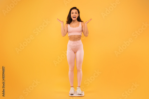 A young woman stands on a scale, expressing surprise and excitement with her hands raised. She is dressed in light-colored active wear and white sneakers, set against a vibrant yellow backdrop.