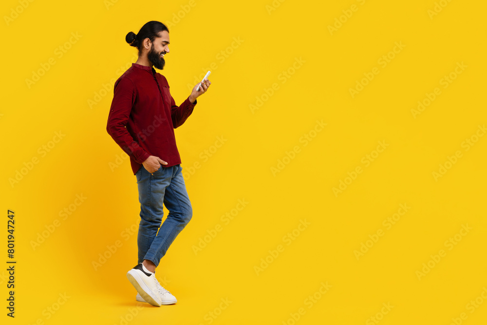Indian man wearing a red sweater is focused on his cell phone screen, scrolling and reading content. He is standing on yellow studio background, copy space Indian man wearing a red sweater is focused on his cell phone screen, scrolling and reading content. He is standing on yellow studio background, copy space