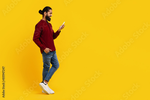 Indian man wearing a red sweater is focused on his cell phone screen, scrolling and reading content. He is standing on yellow studio background, copy space