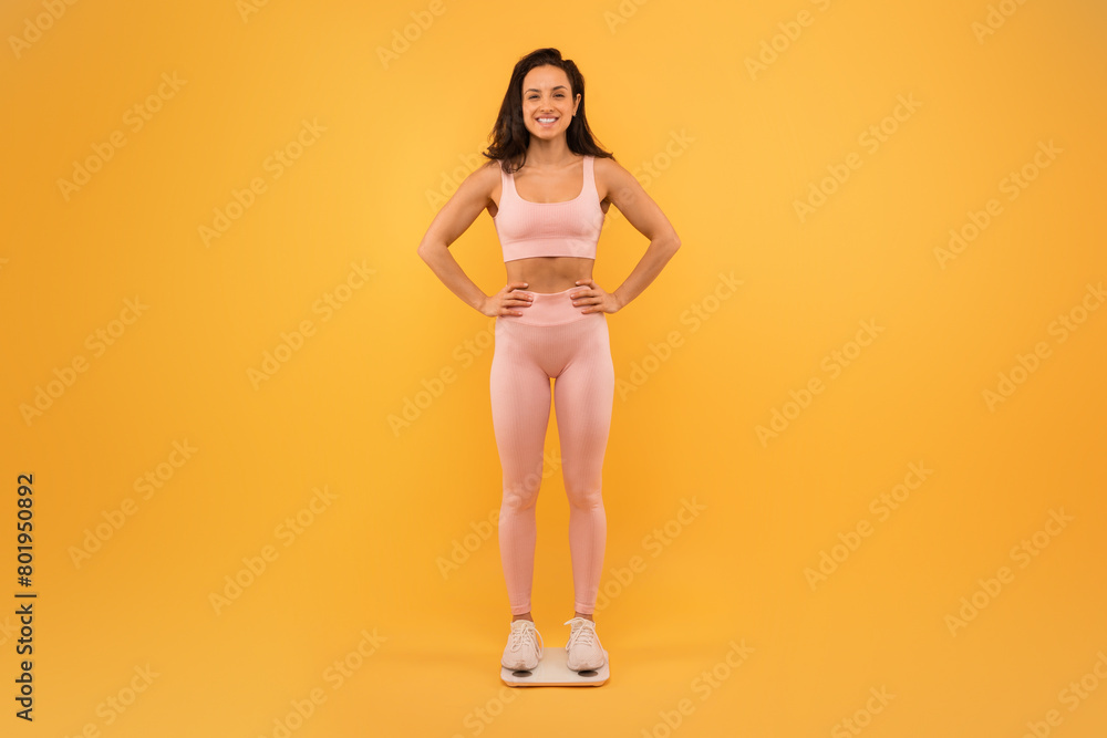 A smiling young woman stands confidently with her hands on her hips, dressed in a light pink sports bra and leggings, checking her weight A smiling young woman stands confidently with her hands on her hips, dressed in a light pink sports bra and leggings, checking her weight
