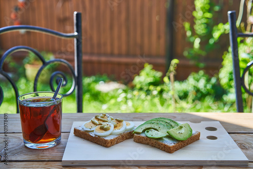 Two open face sandwiches from wholegrain bread spreaded by fresh cheese and decorated with slices of hard boiled egg and fresh avocado on wooden board and cup of black tea served outside as breakfast.
