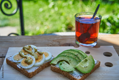 Two wholegrain open sandwiches spreaded by cheese wih slices of fresh avocado and hard boiled egg served with cup of black tea in the home garden as a healthy, light breakfast with balanced nutritions