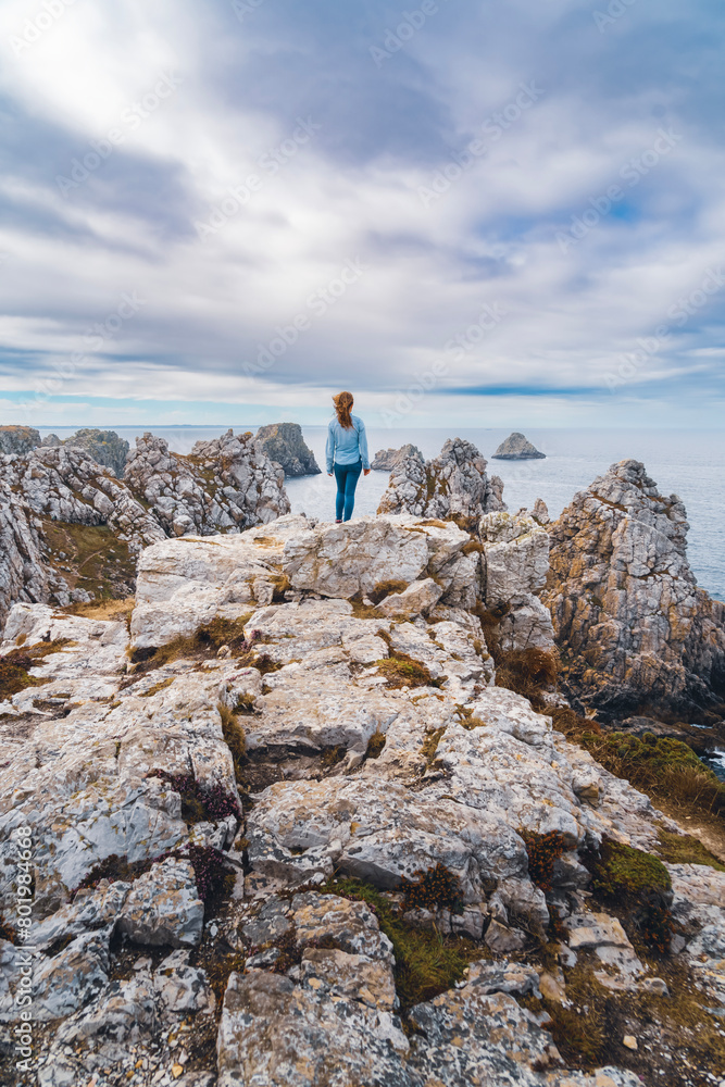 A young girl on vacation looks out over the ocean on top of a rocky promontory in the Pen Hir Point area of the Crozon peninsula, Brittany, France, the three famous islets, France.
