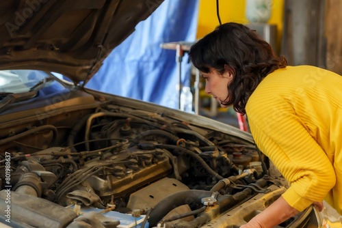 Adult in yellow top inspecting open car hood, examining vehicle engine intently, garage equipment in background, portraying maintenance task. Service work in car repair shop