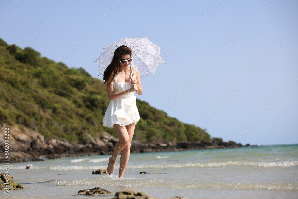 An attractive Asian woman confidently strides along the sandy beach of a tropical island, sheltered from the sun's rays by a white umbrella.