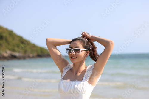 Young attractive ethnic Thai millennial woman posing at suny spreading arms to sides and various postures dressed in white casual clothes stands on the ocean with sandy beach. Love for travel